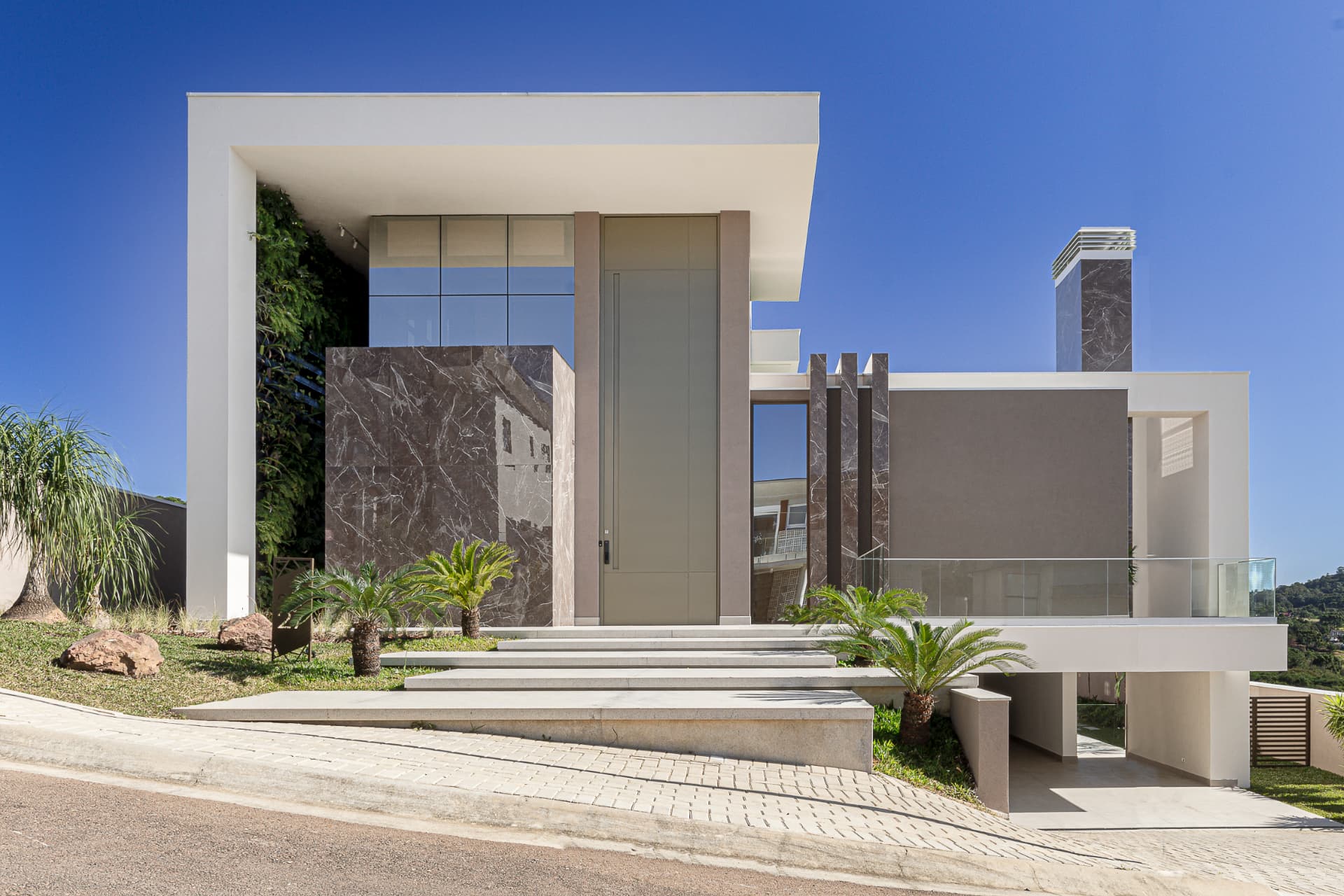 Residência B | T (Casa Pôr-do-Sol) em Curitiba: fachada principal contemporânea com volumes brancos, painéis em mármore escuro, jardim vertical, porta de entrada, escadaria e garagem sob céu azul