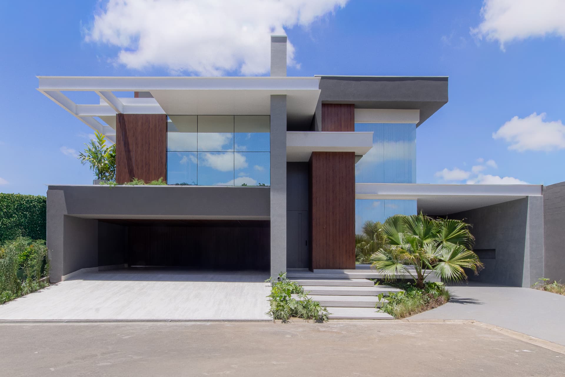 Fachada reformada da Residência C | G com volumes contemporâneos em branco e cinza, esquadrias refletivas, painéis ripados de madeira e garagem integrada ao jardim