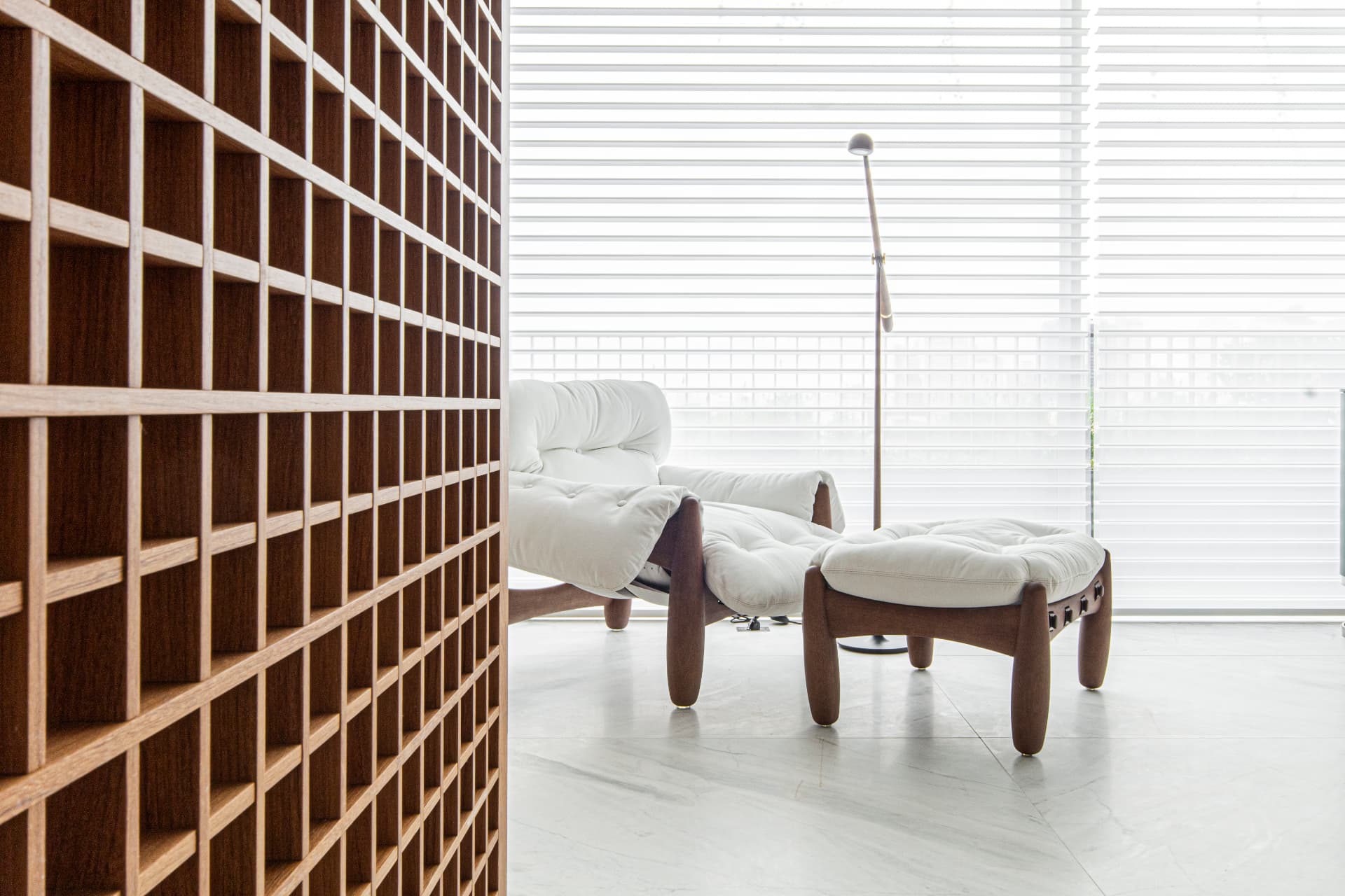 Living do Apartamento B | K em Curitiba com painel vazado de madeira, poltrona branca com ottoman e luz natural filtrada por persianas