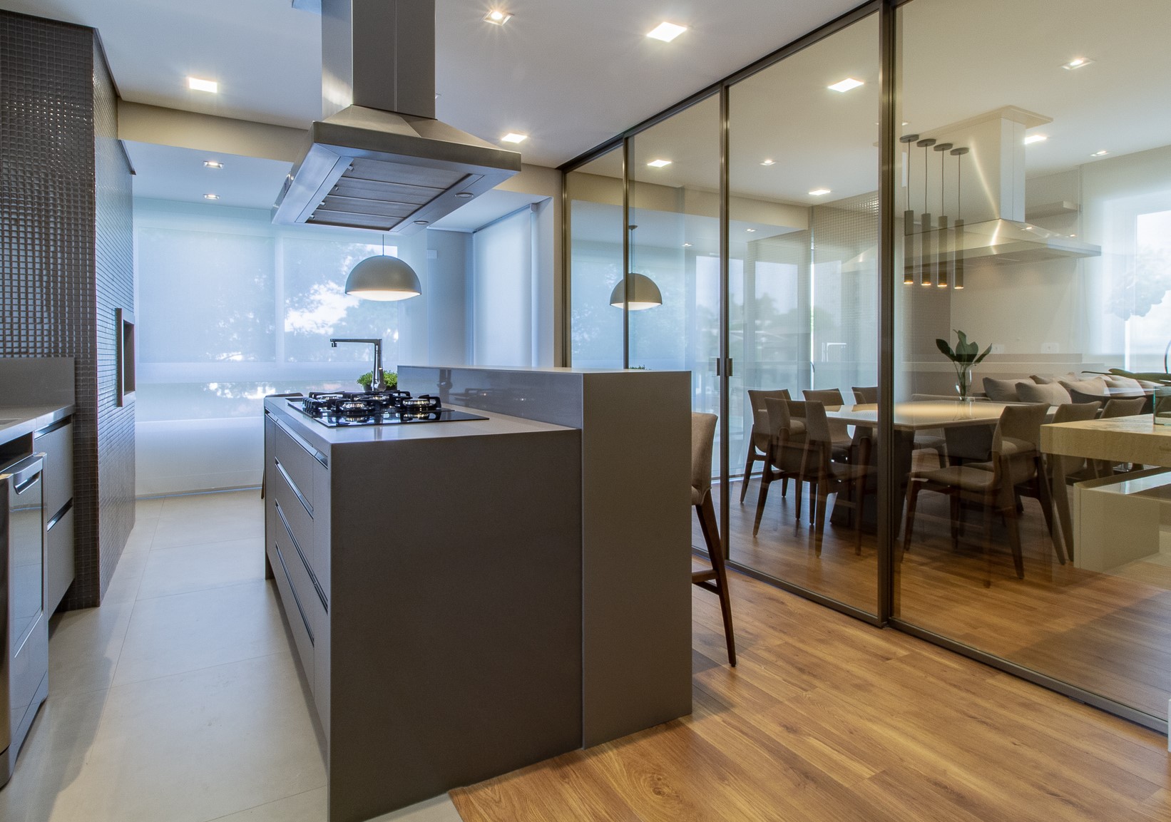 Espaço gourmet integrado à sala de jantar do Apartamento R | S em Curitiba, com ilha central, coifa de aço inox, armários cinza, bancadas claras e divisória de vidro
