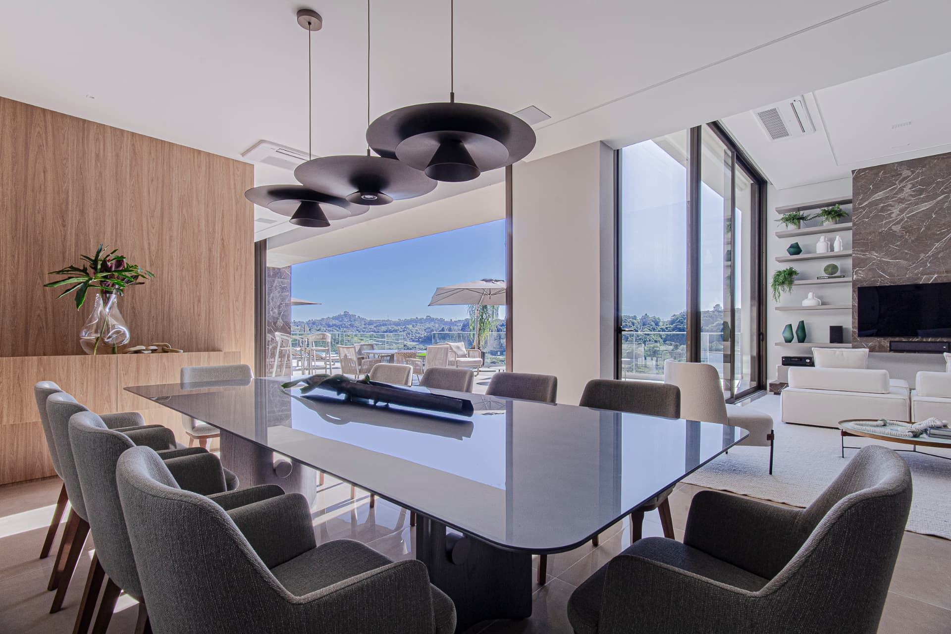 Sala de jantar da Residência B | T (Casa Pôr-do-Sol) em Curitiba: mesa ampla com tampo escuro espelhado, poltronas estofadas cinza, pendentes escuros, painel de madeira e esquadrias amplas com vista para a mata