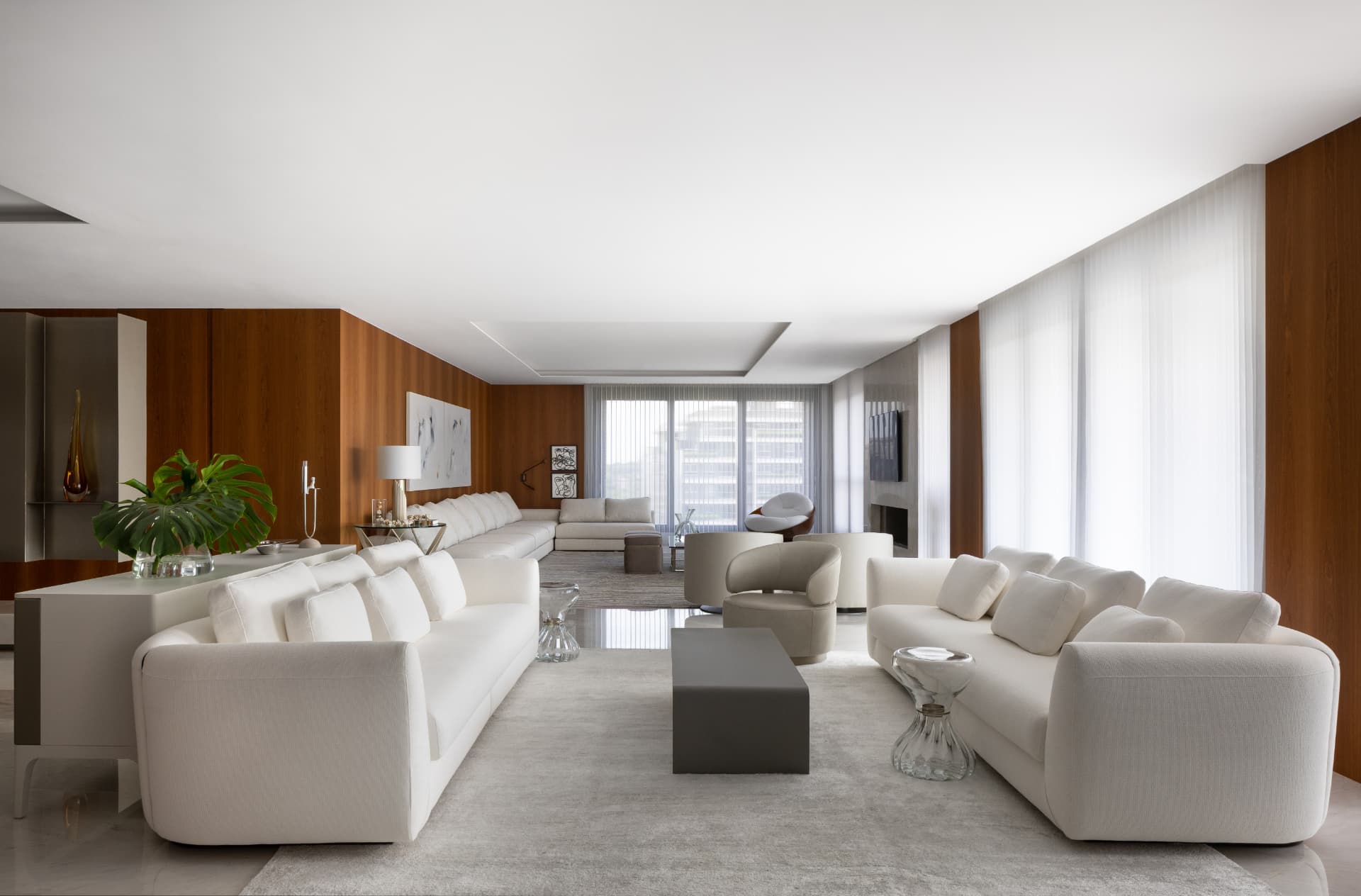 Living amplo do Apartamento V | M com sofás brancos em composição linear, painéis de madeira, poltronas e mesas de centro, luz natural filtrada por cortinas translúcidas e vista para a cidade ao fundo
