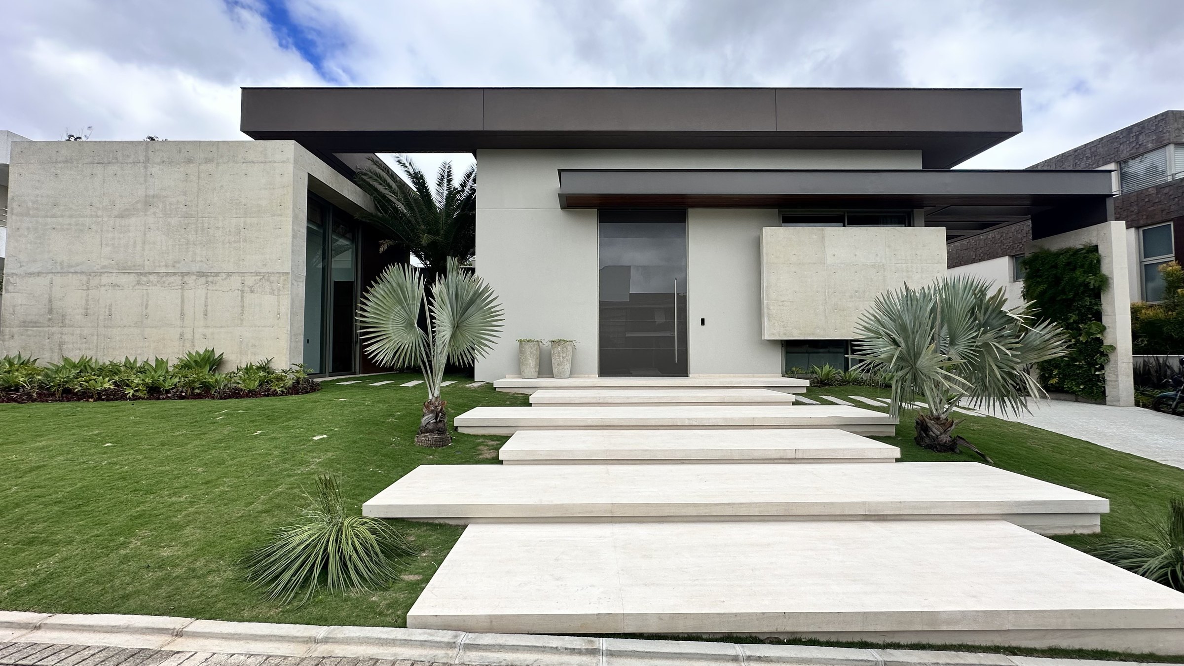 Fachada principal da Residência C | R com volumes em concreto aparente, escadaria monumental, marquise suspensa e paisagismo de palmeiras